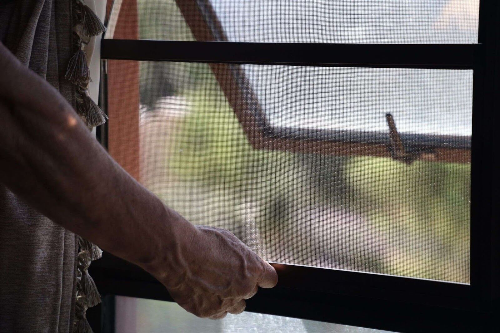 An elderly woman's hands are adjusting an insect screen installed on a dark window frame.
